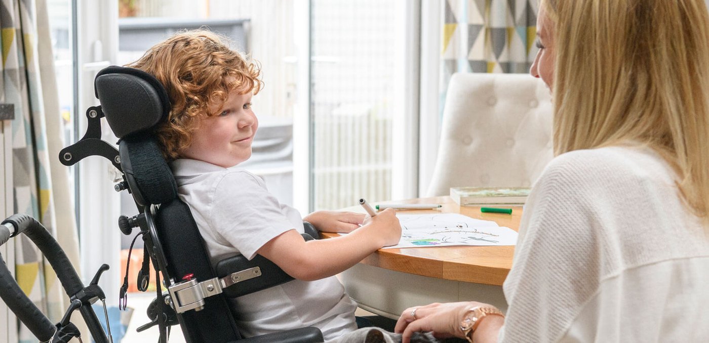 Paediatric & young people's services A boy in a mobility wheelchair writing on a piece of paper with a carer next to him