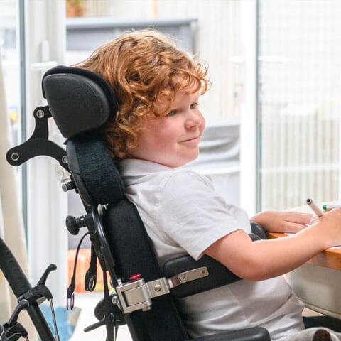 A young boy smiling in a wheelchair at a table writing