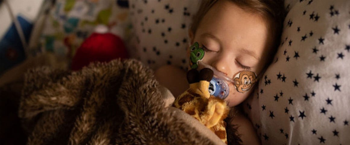 Young girl in hospital bed with oxygen tube cuddling a giraffe teddy